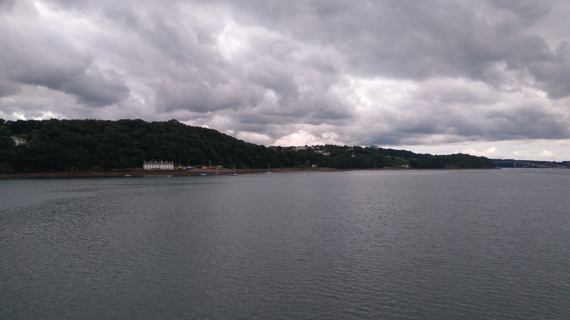 Looking across the Menai Strait at Anglesey on a cloudy summer day. The sea is calm, and the coastline is hilly and wooded.