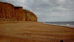 A shingle beach with sandstone cliffs looming up on the left hand side with the sea on the right.