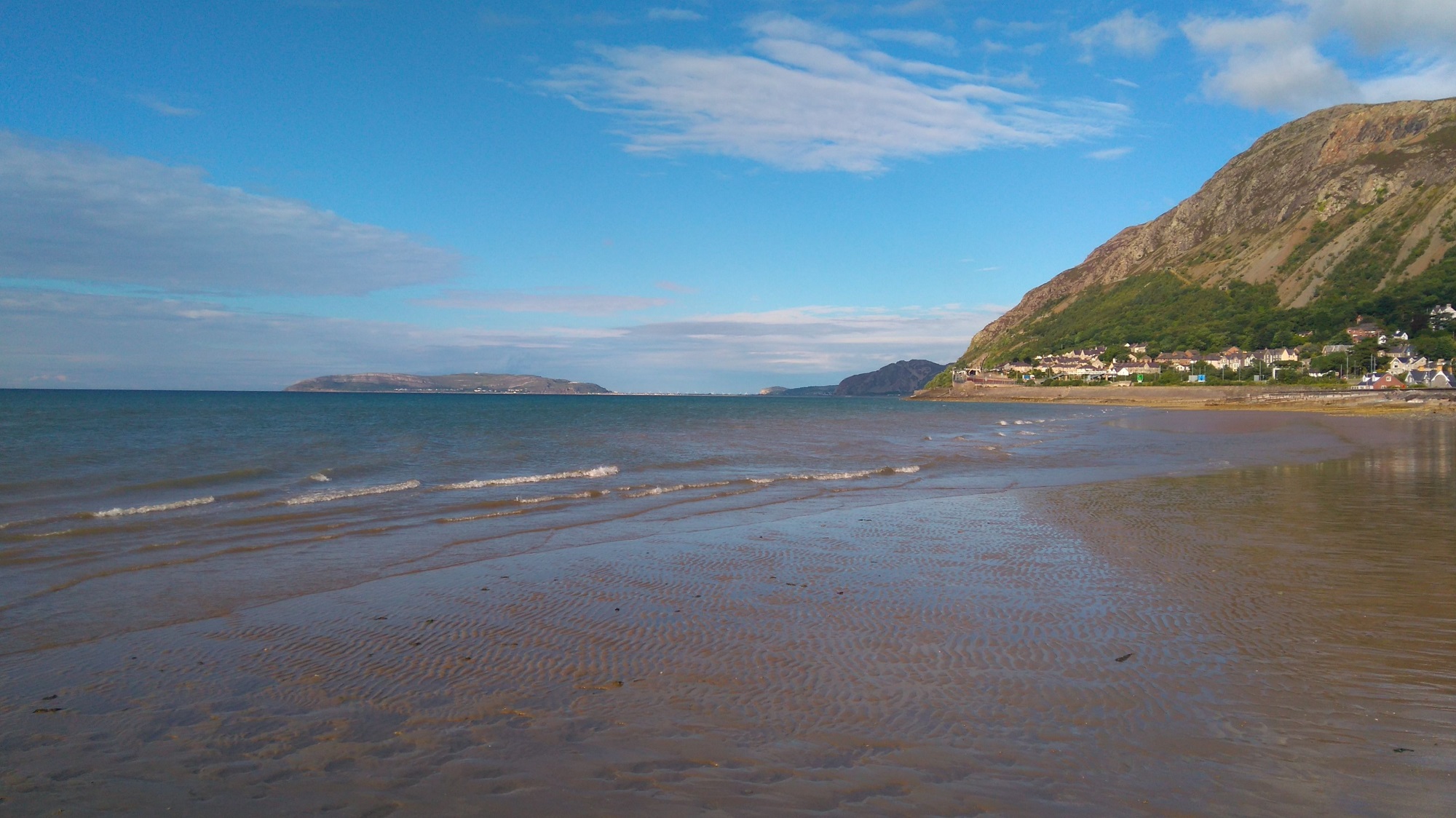 A Welsh mountainside meets the sea under blue skies on a summer day. In the foreground the sea washes across sand flats, while Llandudno is visible in the distance. Llanfairfechan, north Wales.