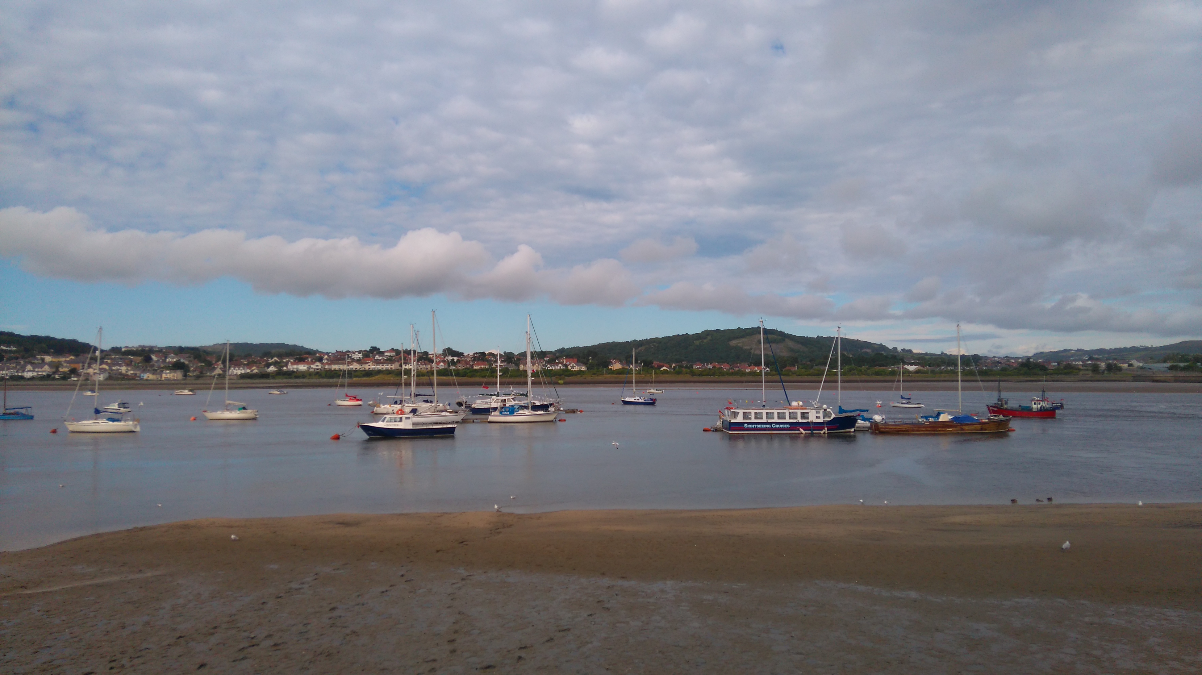 Boats moored to mooring buoys on the River Conwy with mudflats in the foreground. Llandudno Junction is visible in the background.