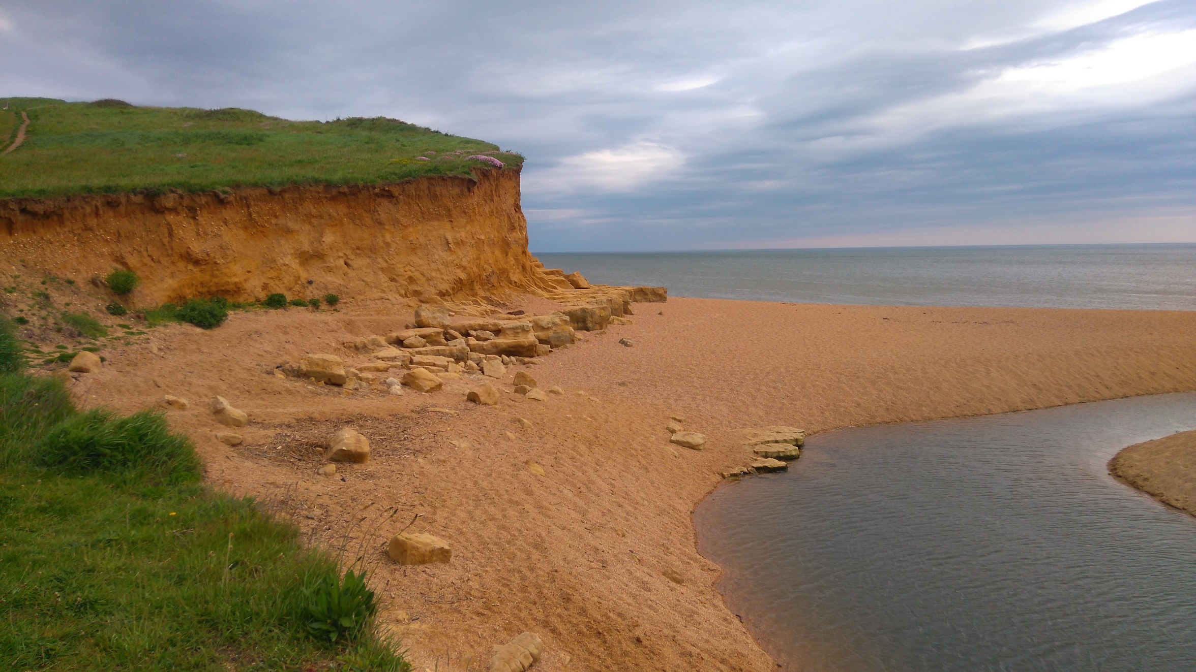 A small cliff by a beach with grassy clifftops under a cloudy sky. The sea is present in the background, while a small waterway is visible on the right.