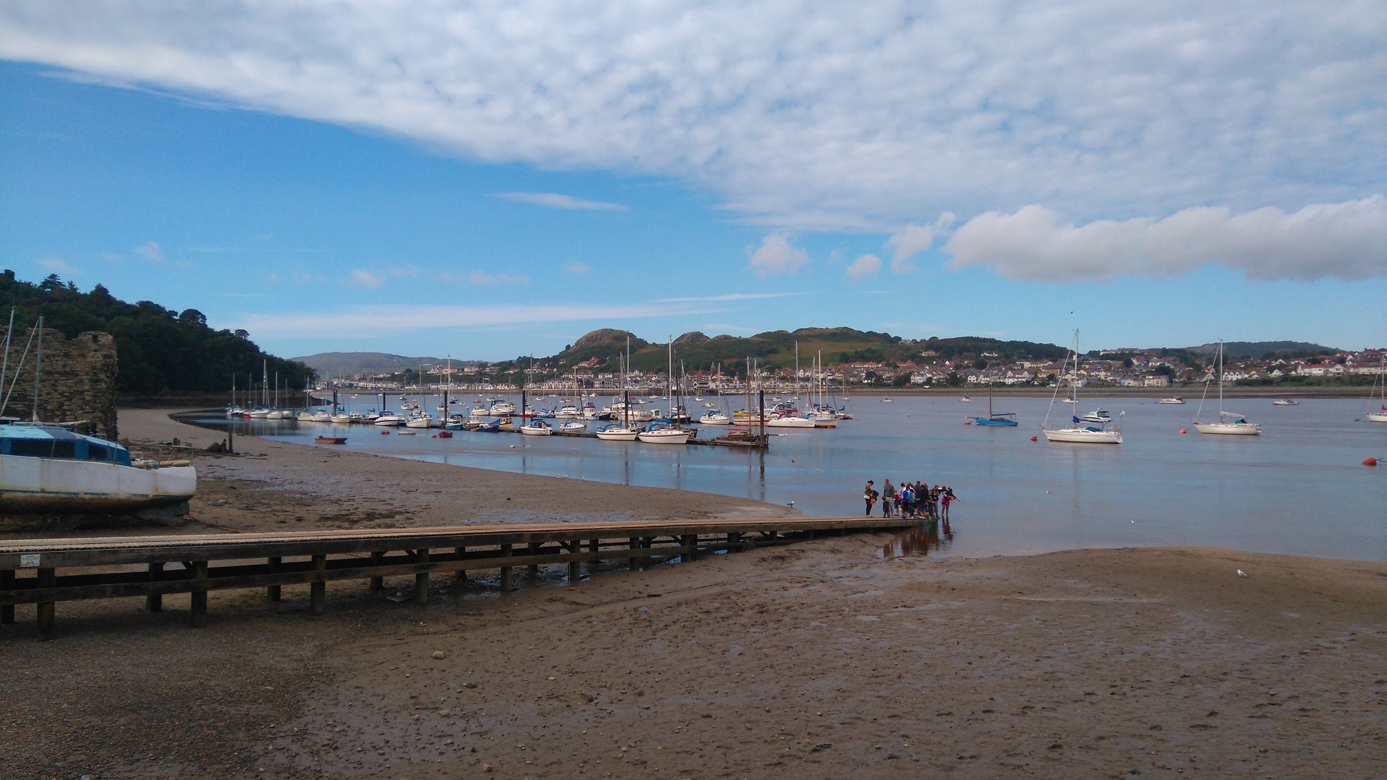 A jetty leads down into the River Conwy where boats are moored to buoys on a summer day. Deganwy is visible across the water.