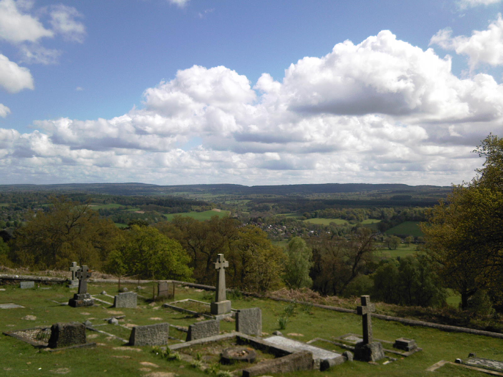 A rural graveyard in the foreground with a vista across the North Downs in Surrey, showing hills, fields and woods under a bright partly cloudy sky.