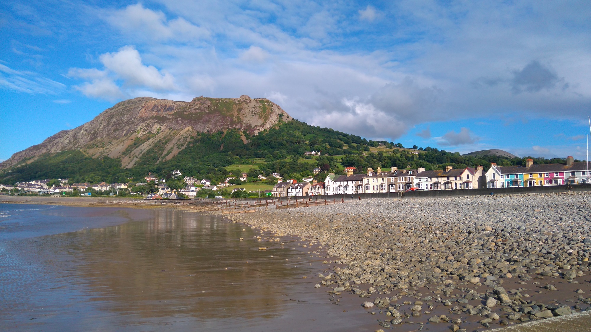 A seaside mountain dominates the beach under a mainly blue sky on a summer day. A row of houses are visible in the middle ground. Llanfairfechan, Wales.