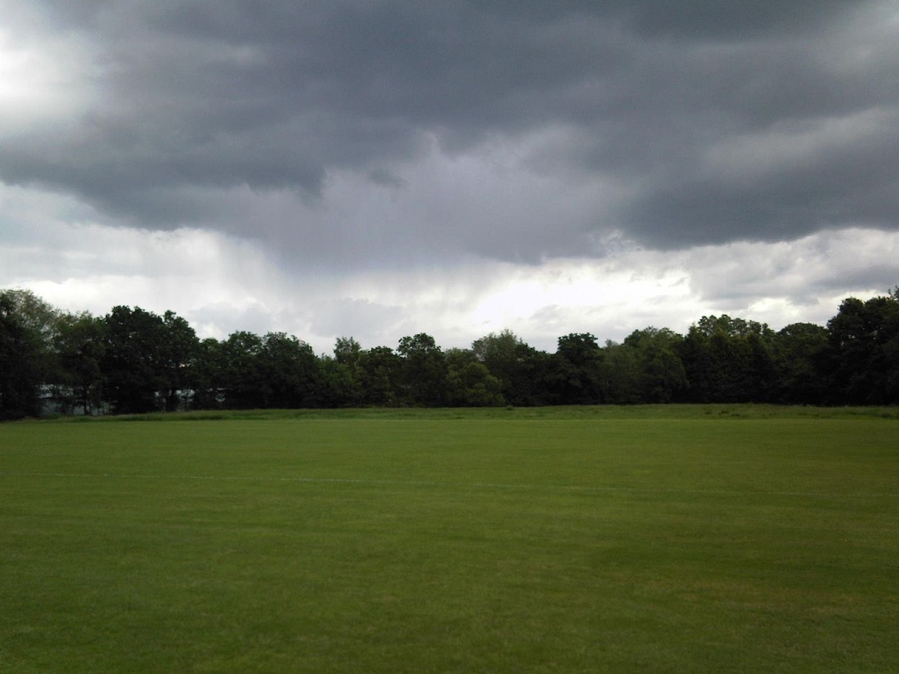 A green playing field lies under a dark grey sky with distant rain falling. The far side of the field is bordered by trees.