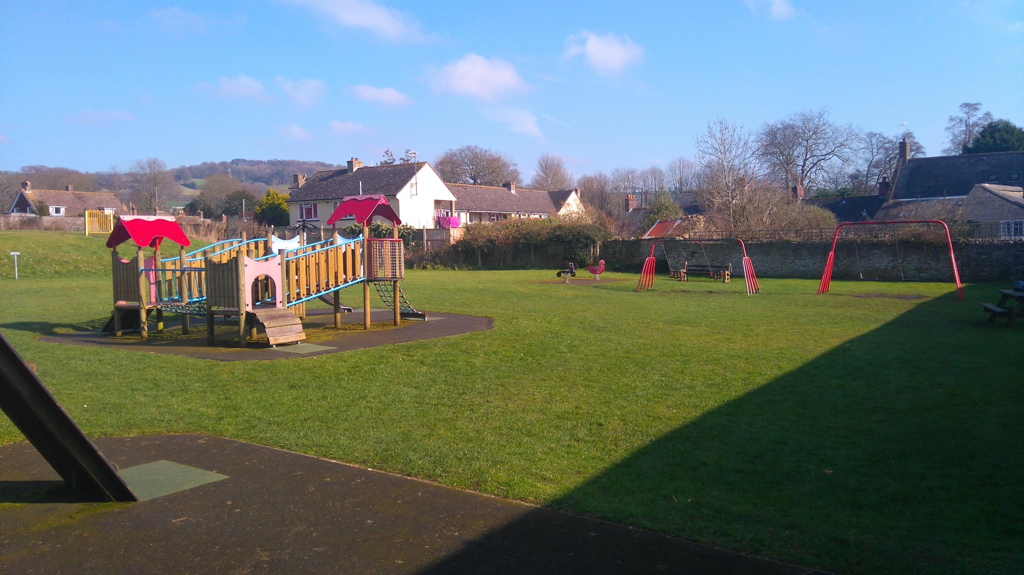 A playpark with climbing area and swings on a sunny day. A rural hill is visible in the background, while houses are present at the far end of the park.