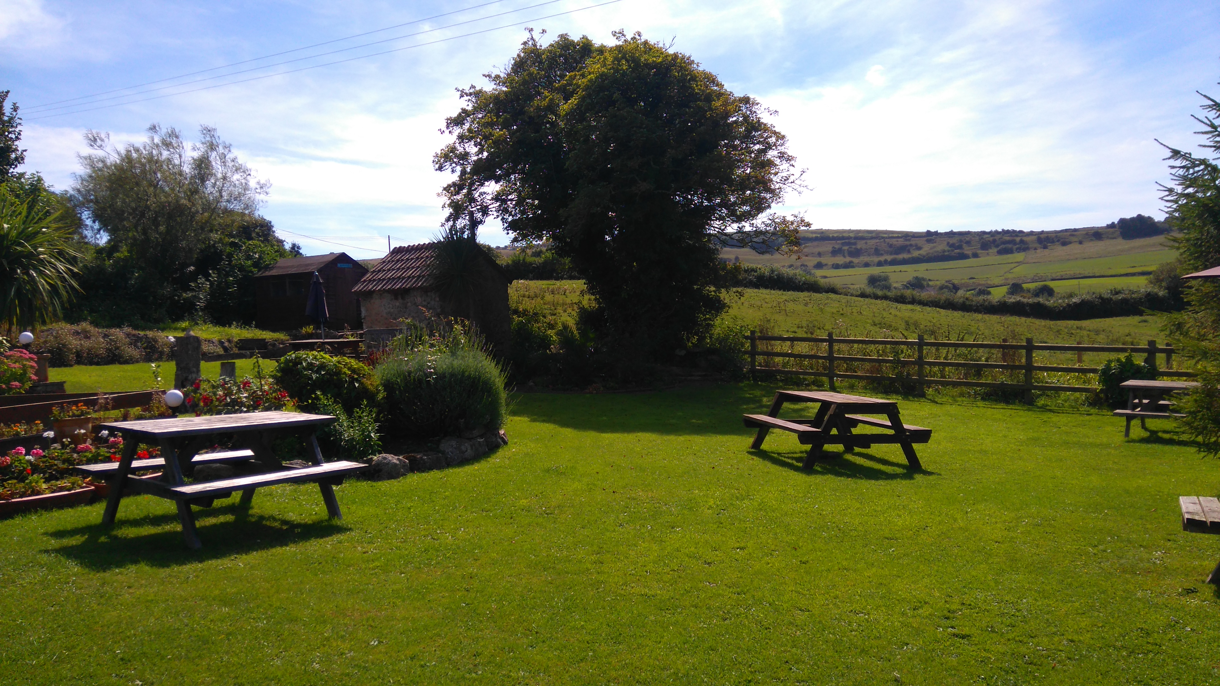 A pub garden in a rural setting on a sunny day. Benches are present within the garden, and fields stretch off into the distance.