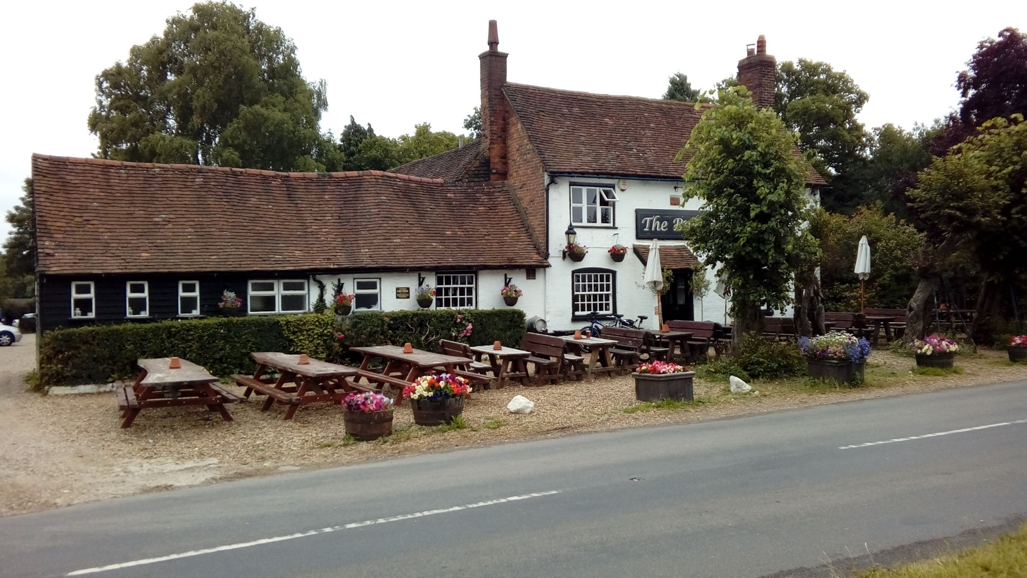 A long pub by a road with a gravel front garden area holding several benches, some large flower pots and a small tree.