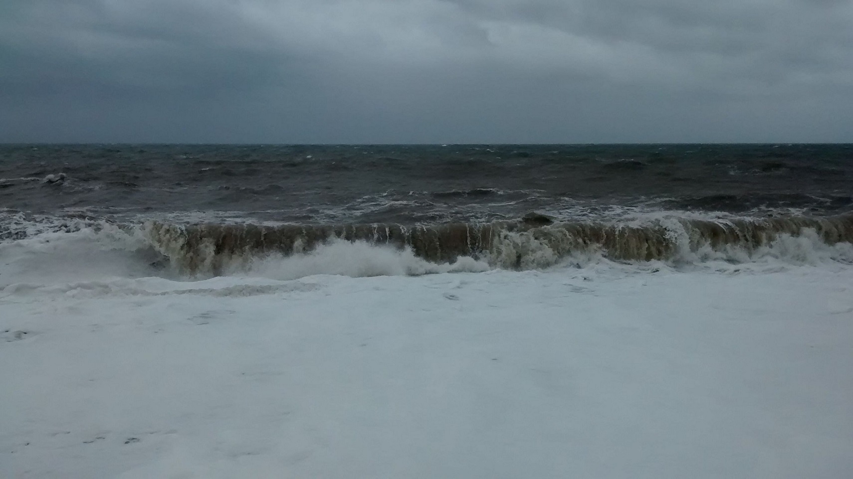 A stormy sea hurls waves against a beach, with white surf run up the beach under a dark cloudy sky.