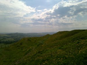 A view across a grassy hillside to a distant rugged rural landscape. Above a bright sky allows shafts of sunlight through the limited cloud cover.