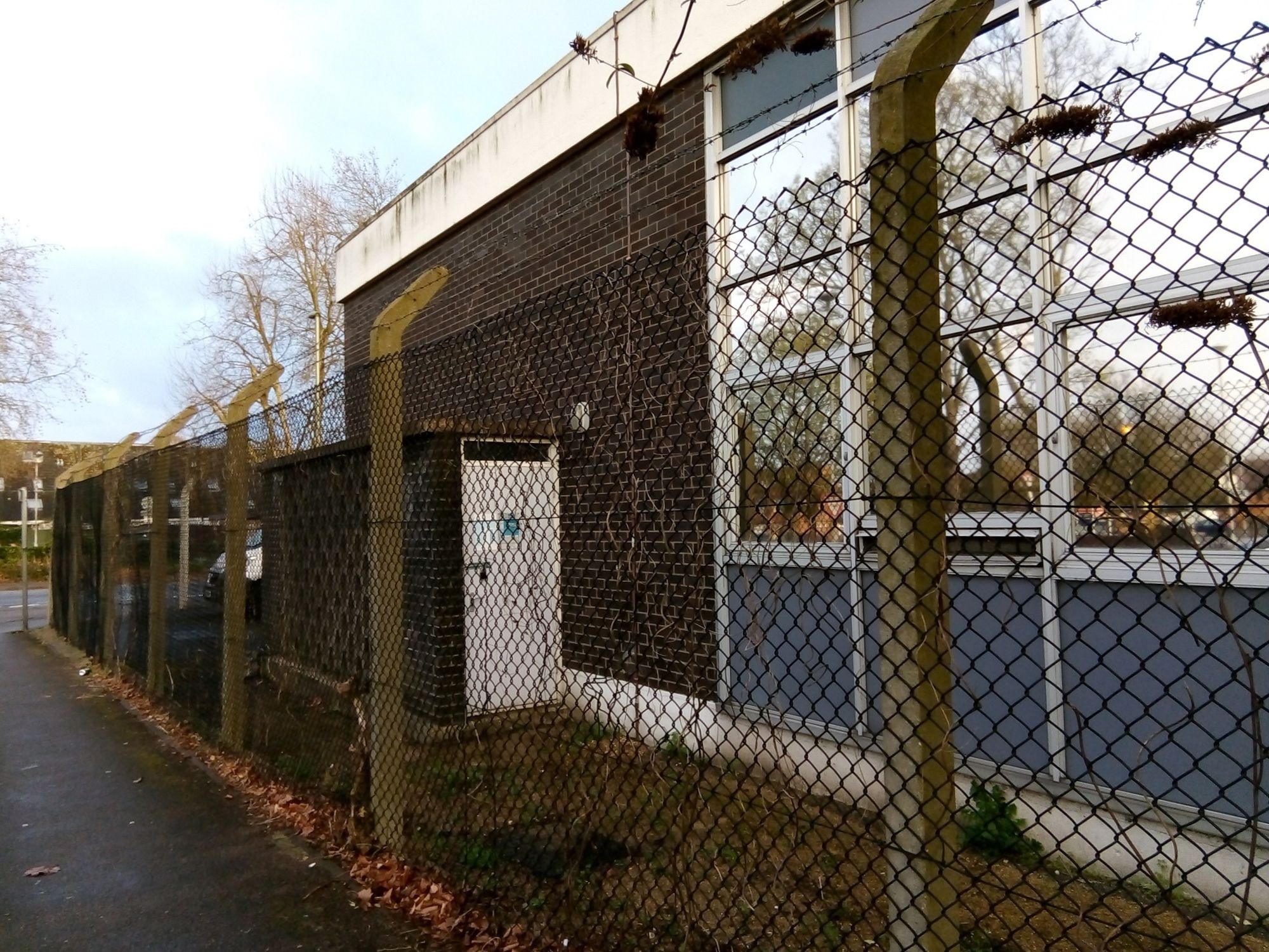 A tall wire fence with concrete posts borders a brick built business property, with paving stones visible behind the fence