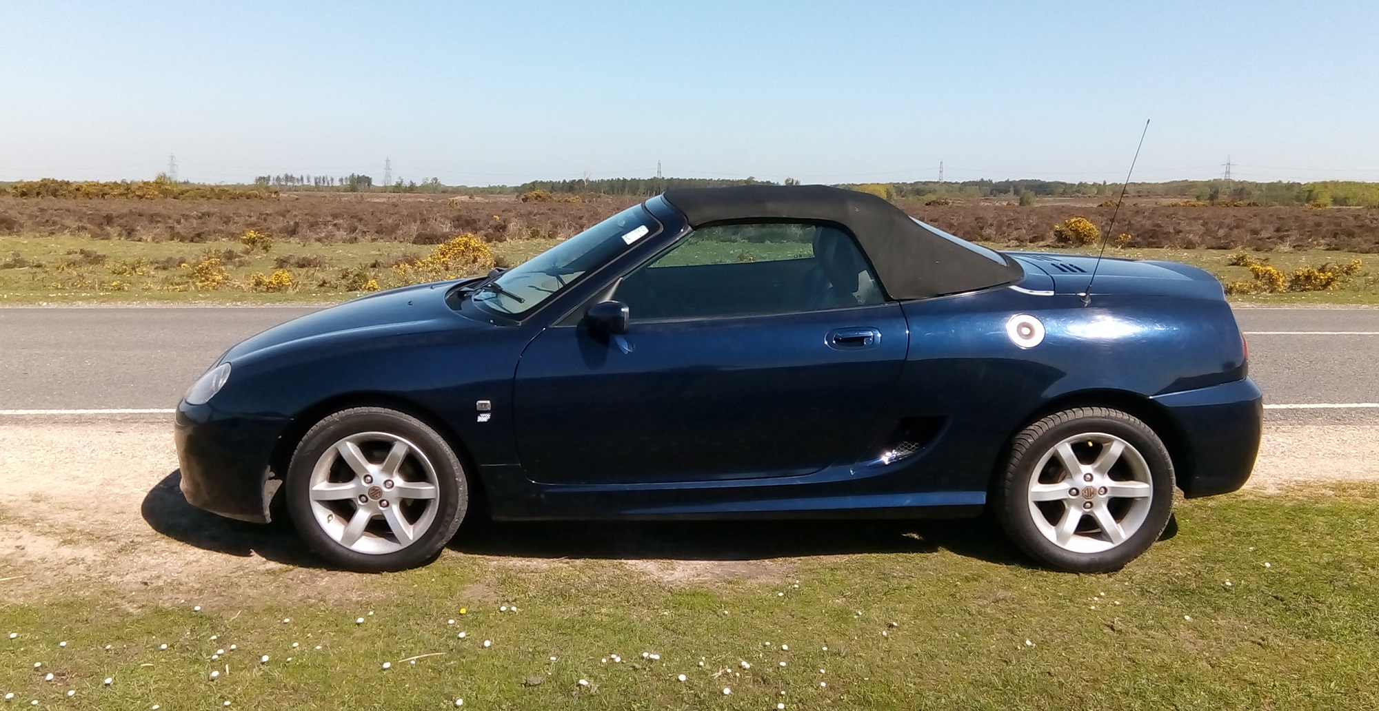 An MG TF dark blue convertible sports car in a sunny rural location.