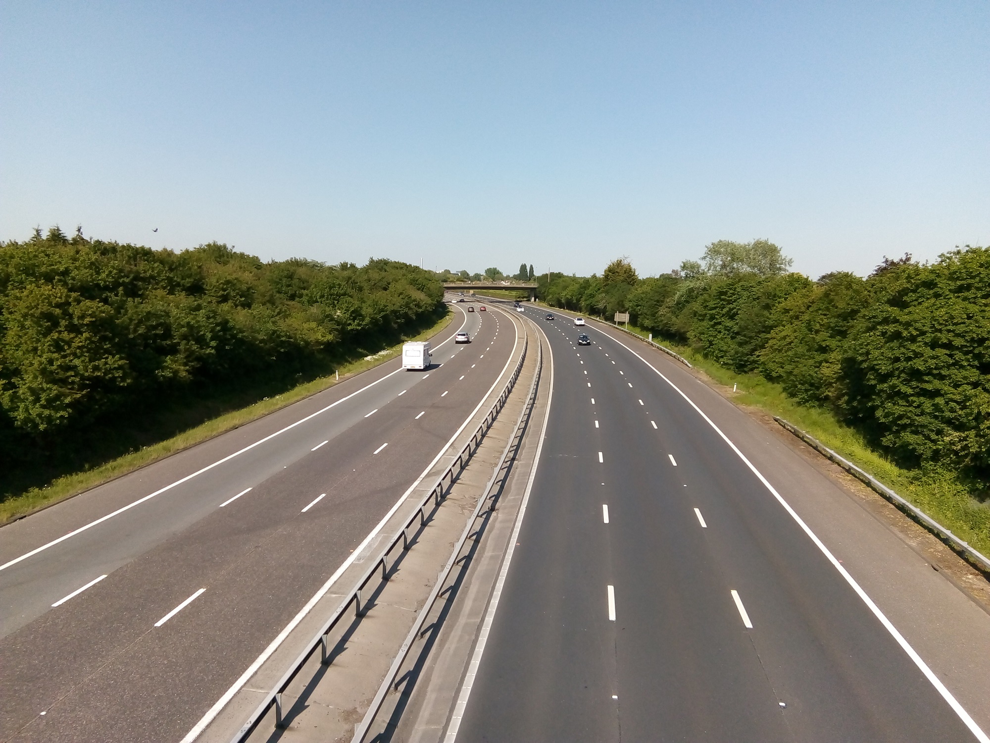 A quiet motorway viewed on a hot sunny afternoon. A bridge is visible in the distance, each side of the motorway is tree lined.