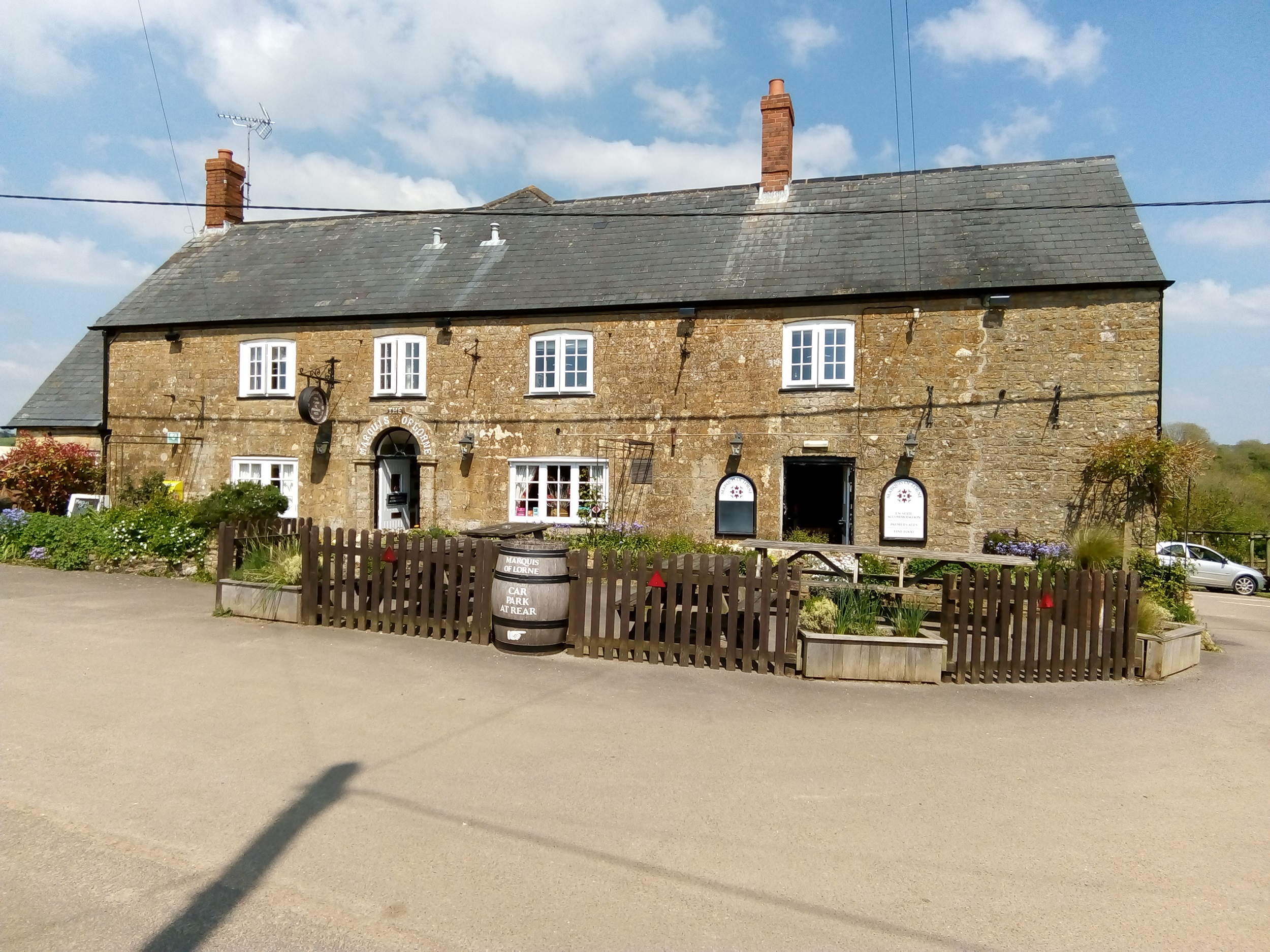 A rural Dorset pub built in stone, and with a dark tiles roof on a sunny day. Seating is visible in the rustic front garden.
