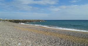 A rocky groyne juts out into the sea by a shingle beach on a summer day under a blue sky holding a few clouds.