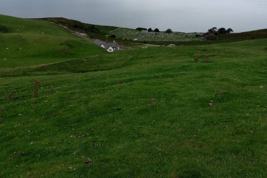 A view of a grassy slope in the foreground towards a grey roofed building and a rural graveyard in the distance against the backdrop of a grey sea