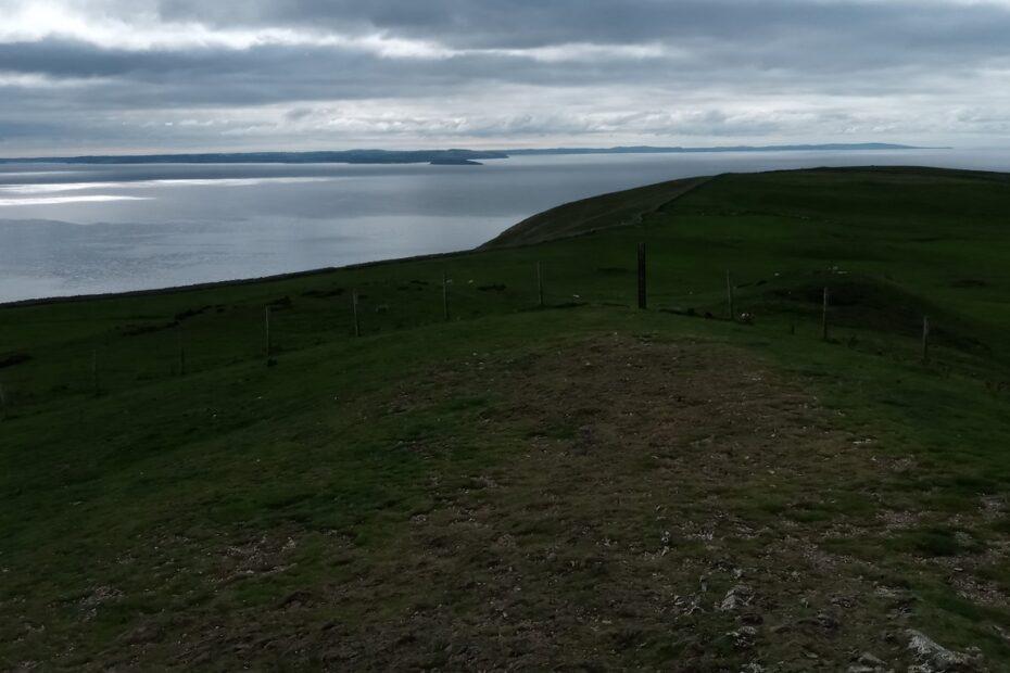 A view across a grassy ridge towards an expanse of sea with a coastline in the distance running left to right under a cloudy sky