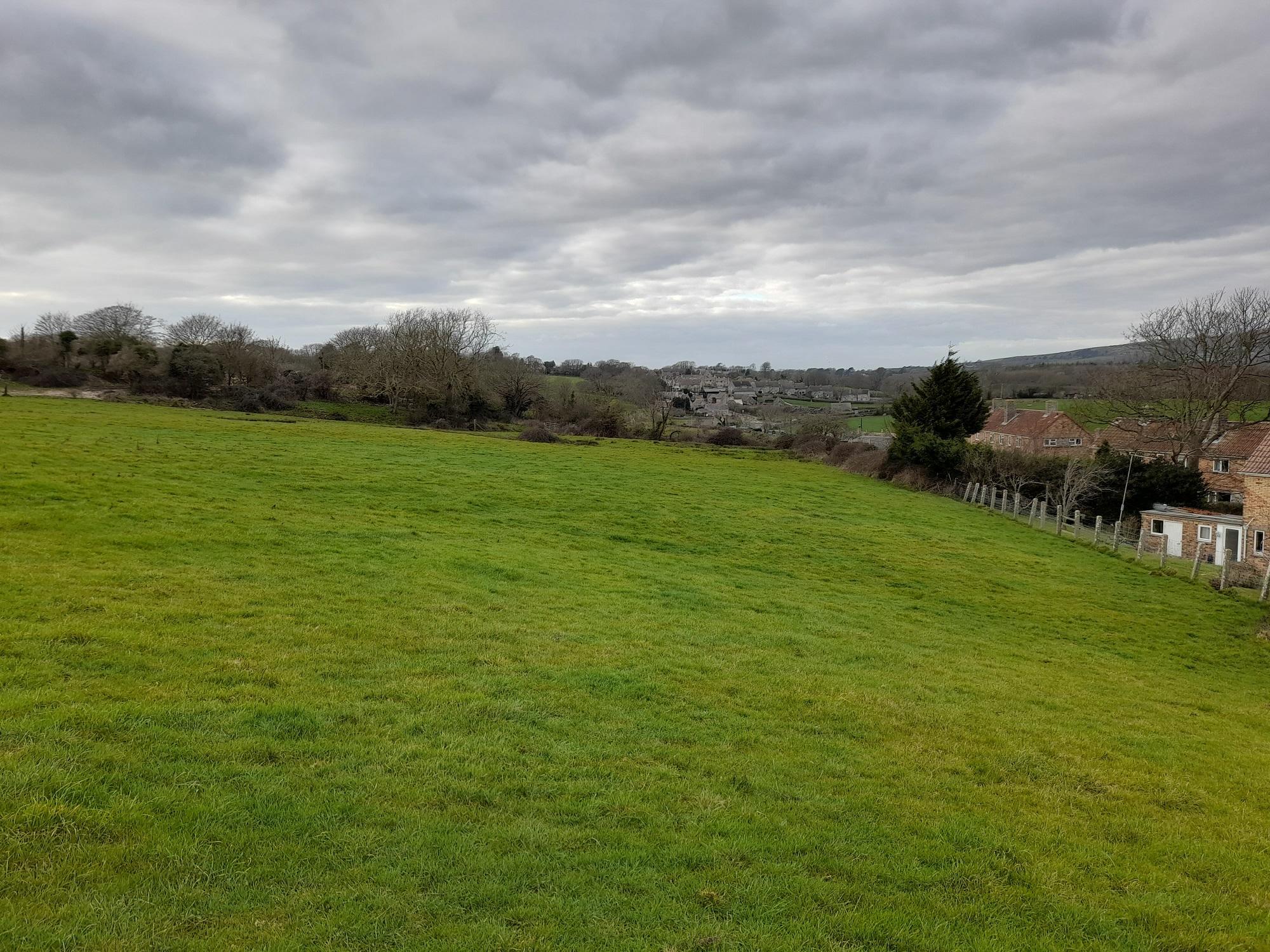 Looking across a large green field on a winter day to the grey stone buildings of Langton Matravers
