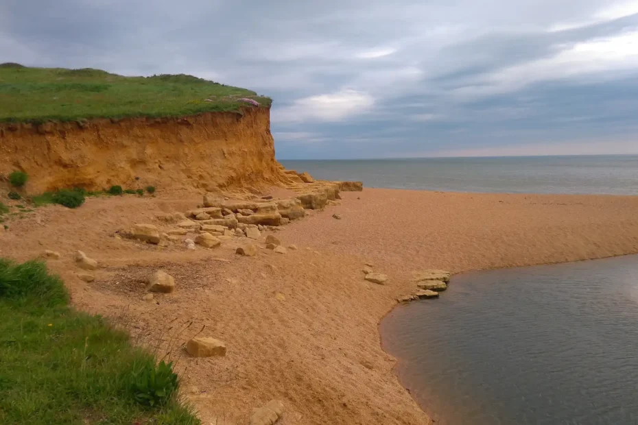A small cliff by a beach with grassy clifftops under a cloudy sky. The sea is present in the background, while a small waterway is visible on the right.